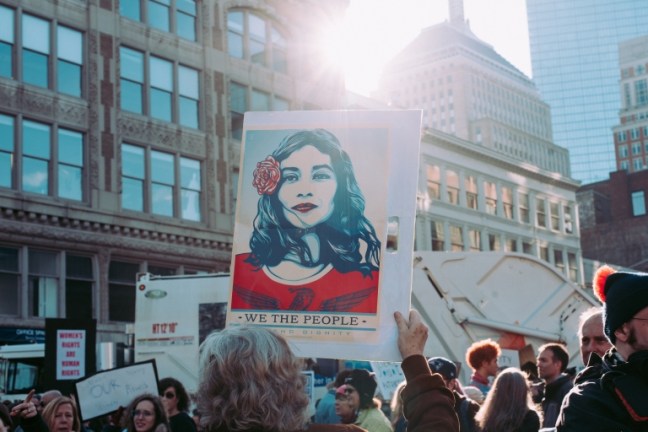 A women's march showing a banner of a beautiful woman with a red rose in her hair and the caption 'We the people'